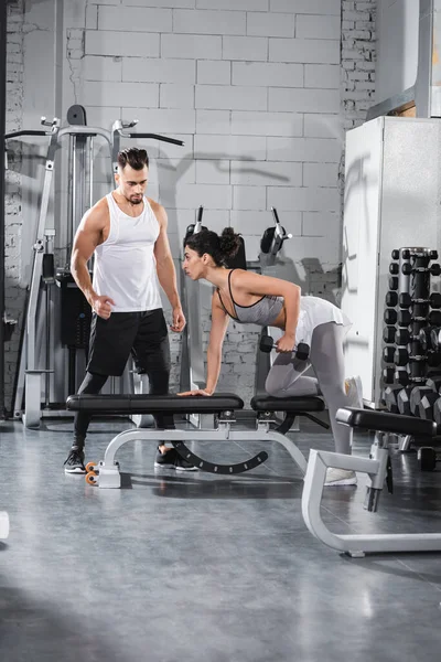 Deportista de Oriente Medio haciendo ejercicio con mancuerna en banco plano cerca de entrenador en el gimnasio - foto de stock