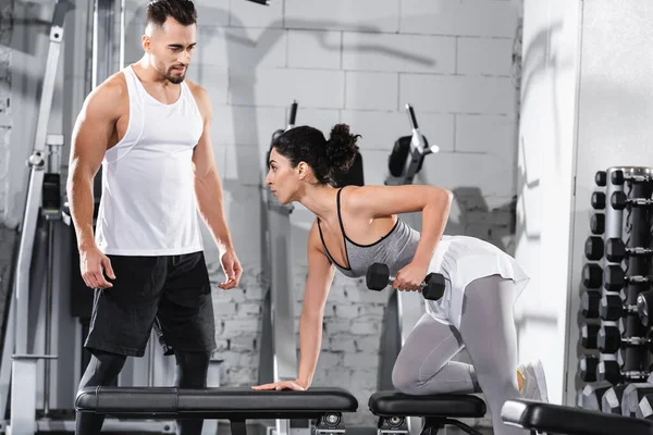 Entrenador de pie cerca de Oriente Medio deportista de entrenamiento con mancuerna en el gimnasio - foto de stock