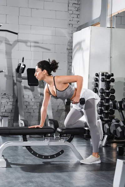 Side view of middle east sportswoman exercising with dumbbell on flat bench in sports center — Stock Photo