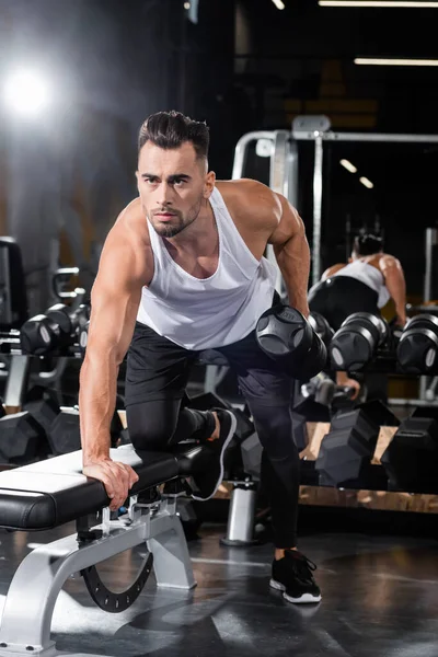 Athletic sportsman working out with dumbbell near flat bench in gym — Stock Photo