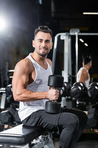 Sportif souriant tenant un haltère assis sur un banc plat dans une salle de gym — Photo de stock