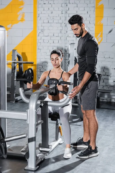 Arabian trainer standing near middle east sportswoman training on ab crunch machine in gym — Stock Photo