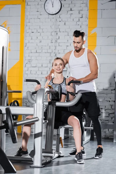 Athletic trainer helping smiling sportswoman near ab crunch machine in sports center — Stock Photo
