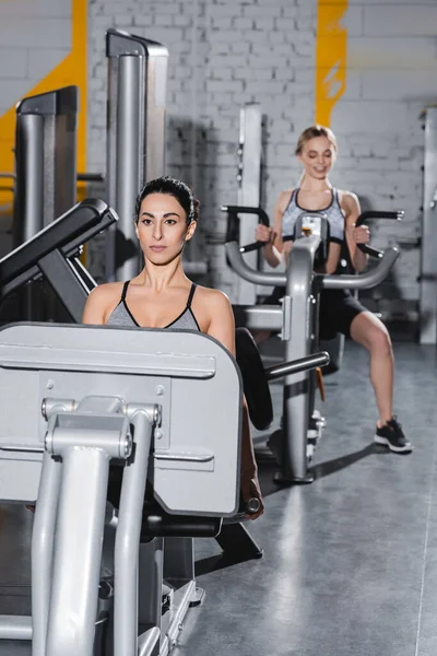 Joven deportista de Oriente Medio haciendo ejercicio en la máquina de prensa de piernas cerca de un amigo borroso en el gimnasio - foto de stock