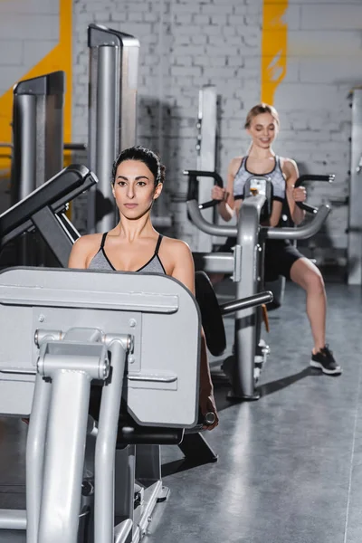 Junge Sportlerin aus dem Mittleren Osten trainiert auf Beinpresse neben verschwommenem Freund im Fitnessstudio — Stockfoto