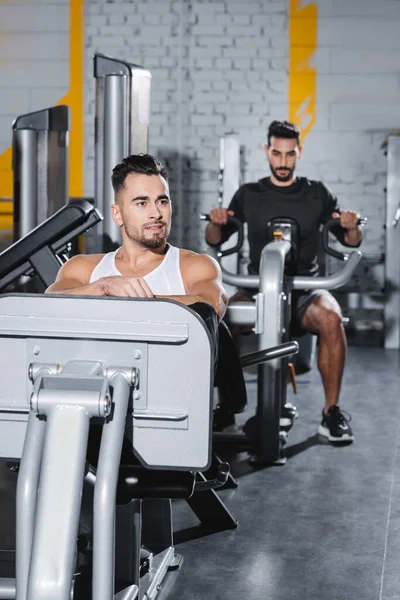 Sorrindo treinamento esportista na perna imprensa máquina perto de amigo árabe borrado no ginásio — Fotografia de Stock