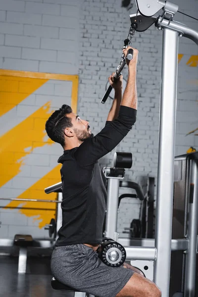 Vista lateral del entrenamiento de deportista árabe con máquina de extracción de lat en el gimnasio - foto de stock