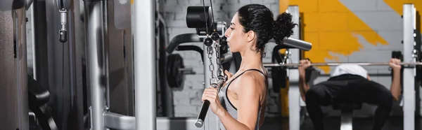 Mujer joven de Oriente Medio entrenando en la máquina de extracción de lat en el centro deportivo, pancarta - foto de stock