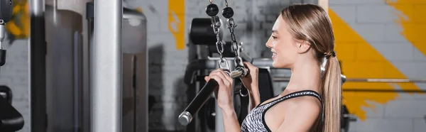 Side view of blonde sportswoman training on lat pulldown machine in gym, banner — Stock Photo