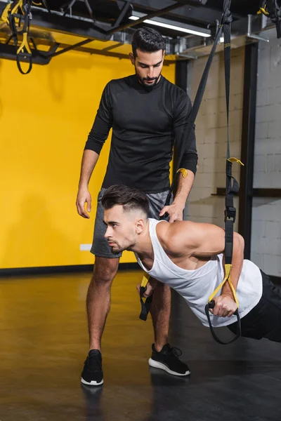 Arabian entrenador de pie cerca de amigo haciendo ejercicio con correas de suspensión en el gimnasio - foto de stock