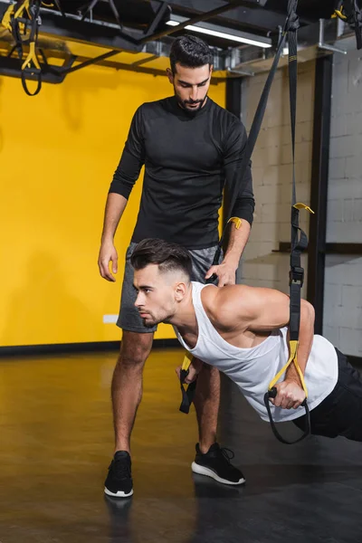 Arabian trainer in piedi vicino amico che lavora con cinghie di sospensione in palestra — Foto stock