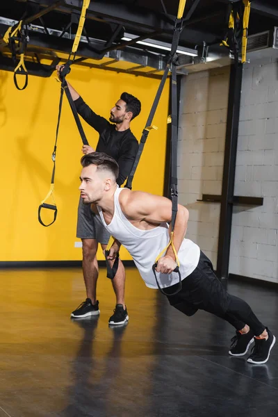 Athletic sportsman pulling suspension straps near muslim friend — Stock Photo