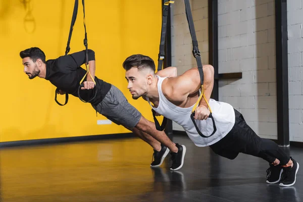 Young multiethnic sportsmen training with suspension straps in gym — Stock Photo