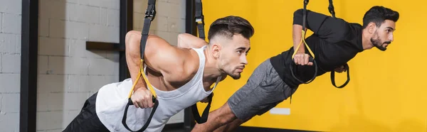 Sportsman training with suspension straps near muslim friend in gym, banner — Stock Photo