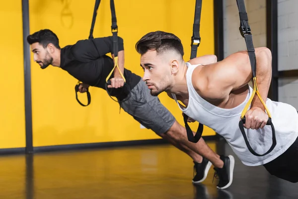 Deportista haciendo ejercicio con correas de suspensión cerca de amigo musulmán borrosa en el gimnasio - foto de stock