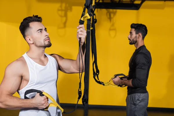 Joven sosteniendo correas de suspensión cerca borrosa amigo árabe en el gimnasio - foto de stock