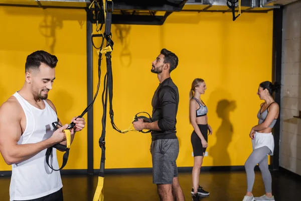 Deportista sonriente sosteniendo correas de suspensión cerca de amigos confusos multiétnicos en el gimnasio - foto de stock