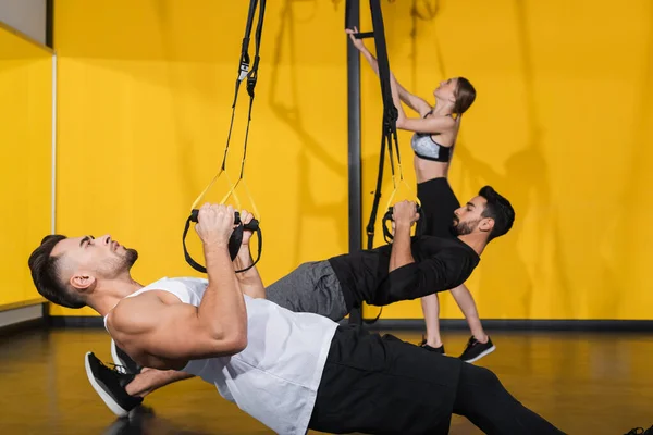 Vista lateral del deportista tirando de correas de suspensión cerca de amigos interracial sobre fondo borroso en el gimnasio - foto de stock