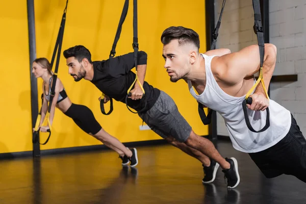 Young sportsman pulling suspension straps near blurred multiethnic friends in gym — Stock Photo