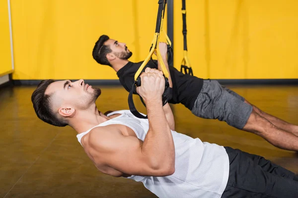 Вид сбоку атлетического спортсмена дергающего ремни подвески рядом с размытым арабским другом тренирующимся в спортзале — стоковое фото