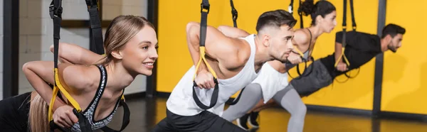 Blonde sportswoman working out with suspension straps near blurred people in gym, banner — Stock Photo