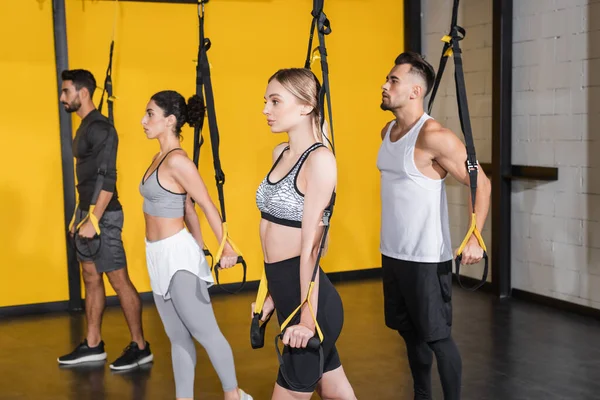 Sportswoman pulling suspension straps while training with interracial friends in sports center — Stock Photo