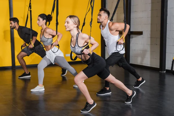 Deportiva positiva tirando de correas de suspensión cerca de personas multiétnicas en el gimnasio - foto de stock