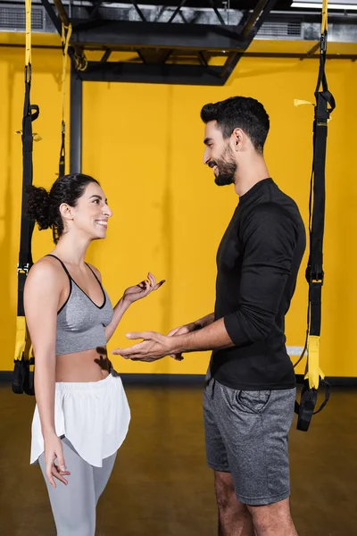 Smiling arabian sportsman talking to middle east woman near suspension straps in gym — Stock Photo
