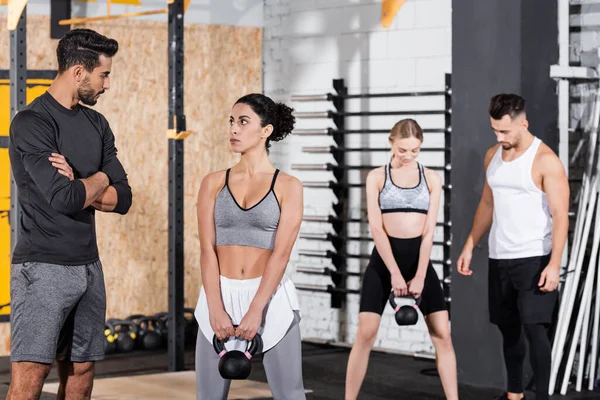 Deportista de Oriente Medio sosteniendo kettlebell cerca entrenador árabe en el gimnasio - foto de stock