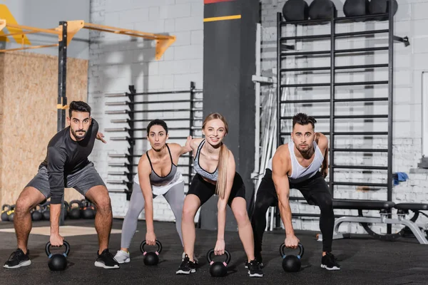 Blonde sportswoman smiling at camera near interracial friends training with kettlebells in gym — Stock Photo
