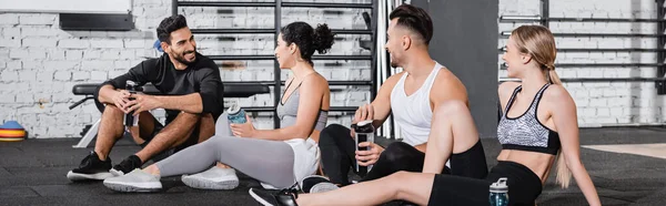 Amigos multiétnicos positivos sosteniendo botellas deportivas en el gimnasio, pancarta - foto de stock