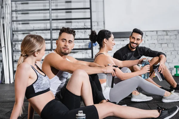 Sportsman holding sports bottle and talking to friend near interracial people in gym — Stock Photo