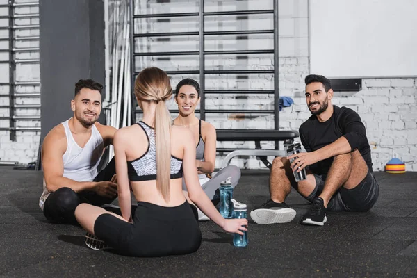 Jóvenes sonrientes multiétnicos con botellas deportivas hablando con un amigo en el gimnasio - foto de stock