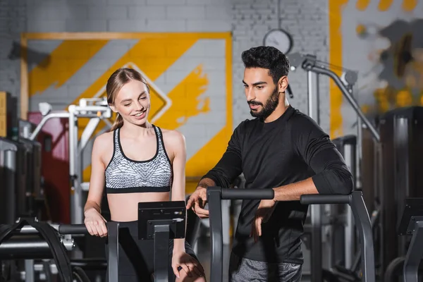 Entraînement sportif positif sur l'entraîneur elliptique près de l'entraîneur arabe dans la salle de gym — Photo de stock