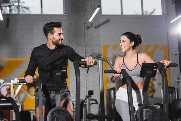 Sonriente deportista árabe mirando a la deportista de Oriente Medio en elíptica en el gimnasio - foto de stock