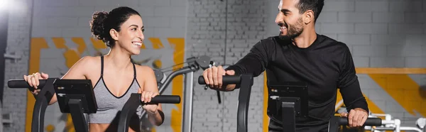 Positive middle east friends training on elliptical trainers in gym, banner — Stock Photo