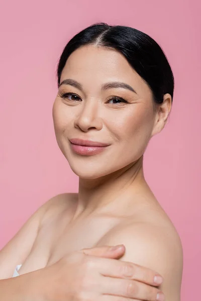 Retrato de mujer asiática sonriente con hombros desnudos mirando a la cámara aislada en rosa - foto de stock