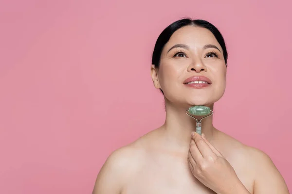 Asian woman with naked shoulders massaging face with jade roller isolated on pink — Stock Photo