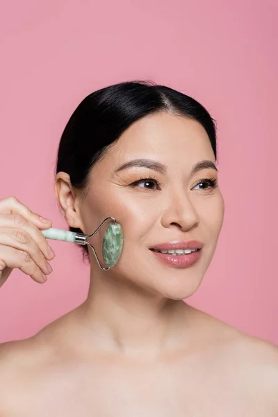 Positive asiatique femme massage visage avec jade rouleau isolé sur rose — Photo de stock
