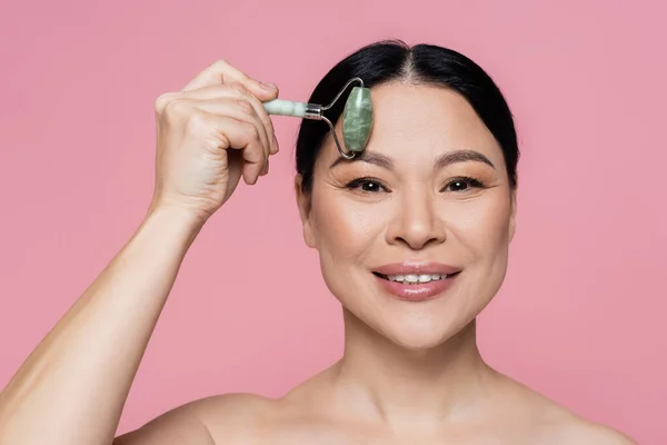 Smiling asian woman with naked shoulders massaging forehead with jade roller isolated on pink — Stock Photo