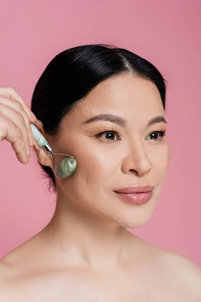 Asian woman with naked shoulders massaging cheek with jade roller isolated on pink — Stock Photo