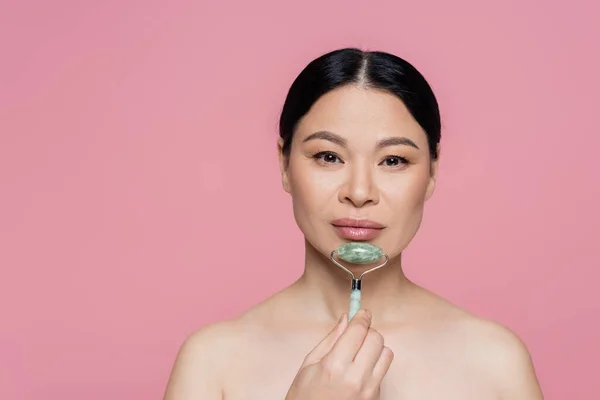 Asian woman with naked shoulders massaging chin with jade roller isolated on pink — Stock Photo