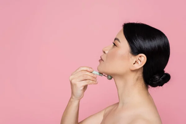 Side view of asian woman with naked shoulders massaging neck with jade roller isolated on pink — Stock Photo