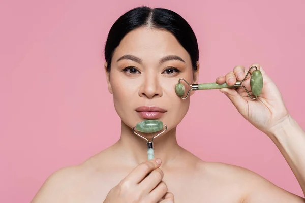 Asiatische Frau massiert Gesicht mit Jade-Rollen isoliert auf rosa — Stockfoto