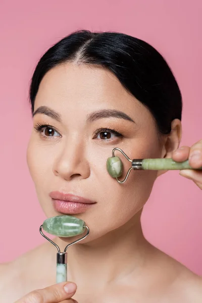 Asian woman with naked shoulders massaging face with jade rollers isolated on pink — Stock Photo