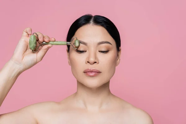 Asian woman with naked shoulders massaging eyebrow with jade roller isolated on pink — Stock Photo