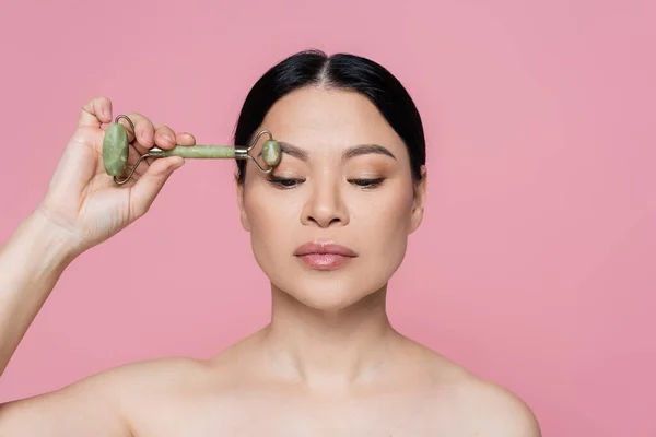 Femme asiatique aux épaules nues massant les sourcils avec rouleau de jade isolé sur rose — Photo de stock