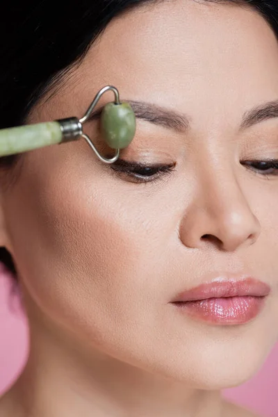 Vue rapprochée de la femme asiatique massant le visage avec un rouleau de jade flou isolé sur rose — Photo de stock