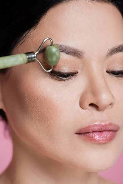 Close up view of asian woman massaging face with blurred jade roller isolated on pink — Stock Photo