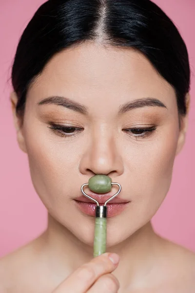 Asian woman massaging face with jade roller isolated on pink — Stock Photo
